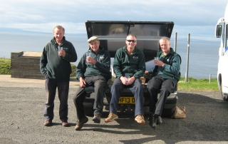 4 men in green jackets sat in the back of a vehicle, holding drinks.